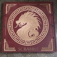 A square wooden Scrabble board with a dark reddish-brown finish and gold laser-engraved details, including a central dragon motif and the word 'SCRABBLE' at the bottom, placed on a patterned black and gold surface.