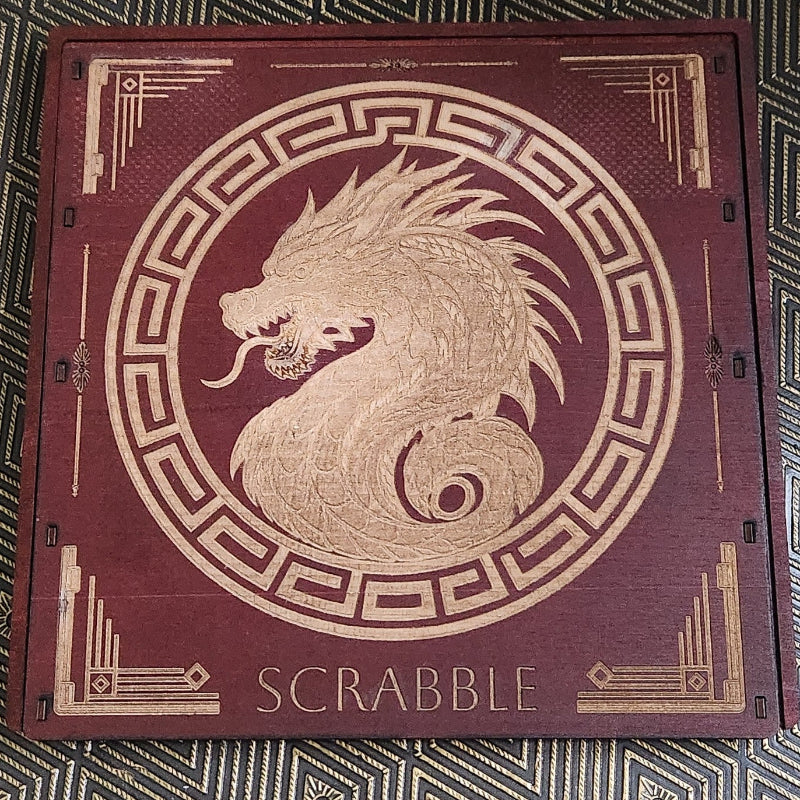 A square wooden Scrabble board with a dark reddish-brown finish and gold laser-engraved details, including a central dragon motif and the word 'SCRABBLE' at the bottom, placed on a patterned black and gold surface.