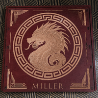 A square wooden box lid featuring an engraved light wood dragon profile inside a circular frame, with geometric corner details and the name 'MILLER' at the bottom, placed on a patterned dark surface.