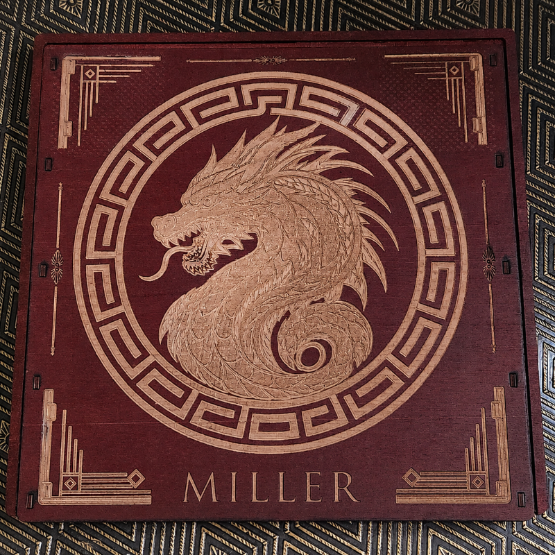 A square wooden box lid featuring an engraved light wood dragon profile inside a circular frame, with geometric corner details and the name 'MILLER' at the bottom, placed on a patterned dark surface.