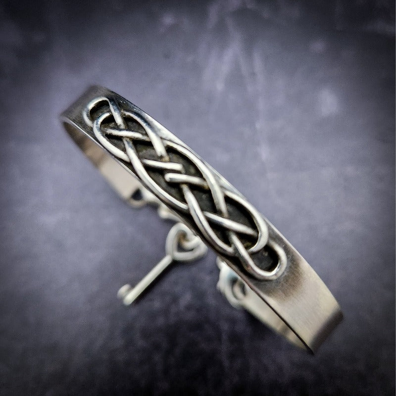 The Cadman Key Keeper Cuff features a chain with a detachable clasp that holds the key to the submissive's lock. With these unique jewelry clasps, the key chain removal is quick and convenient - the key chain can even be linked to a collar during play. Crafted from sterling silver, these luxurious pieces add a sophisticated flair to BDSM boudoirs. By My Secret Heart Studios