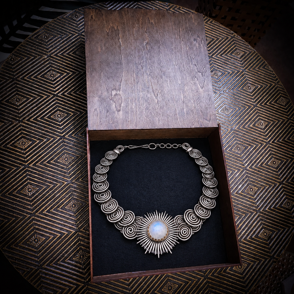 A silver-toned statement necklace with a central sunburst and spiral motifs, displayed inside an open wooden jewelry box on a dark textured surface.