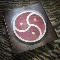A square dark wooden plaque featuring a circular inlaid design with a red and beige S-shape motif and three small gold dots, photographed on a patterned dark background.