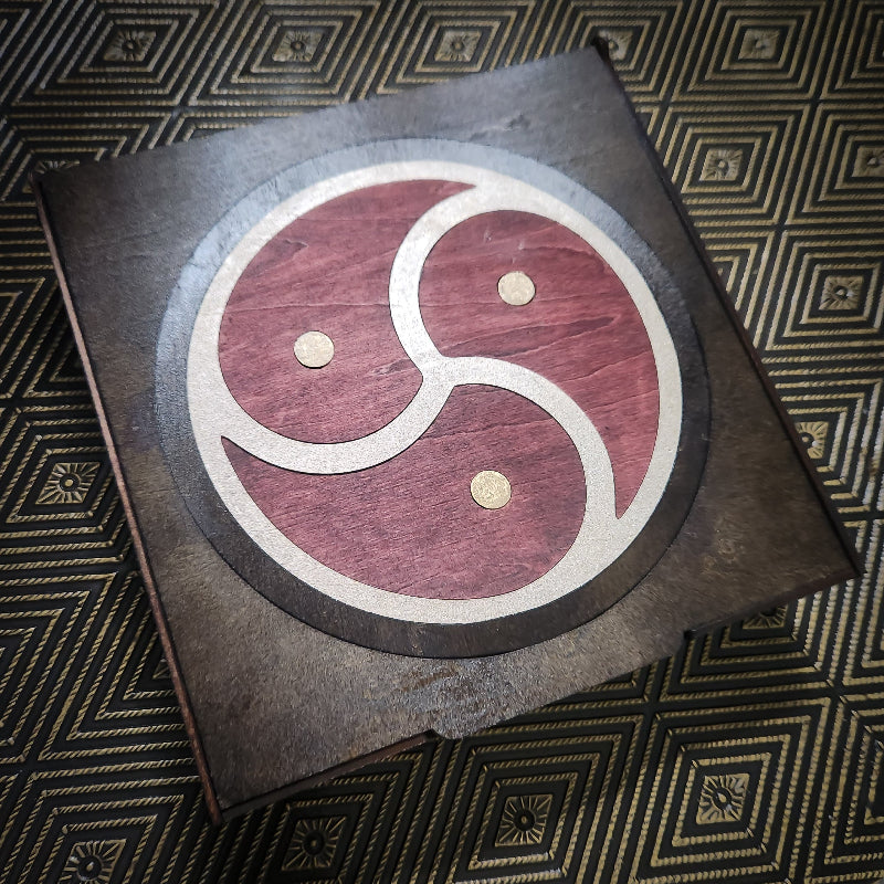 A square dark wooden plaque featuring a circular inlaid design with a red and beige S-shape motif and three small gold dots, photographed on a patterned dark background.