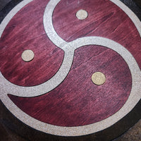 A close-up of a circular wooden plaque or tray featuring a yin-yang style triquetra design in dark reddish-brown wood with a glittery silver border and three small gold-colored circular accents.