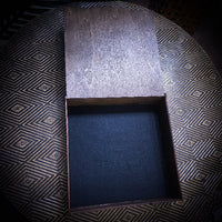 An open rectangular wooden box with a dark textured interior, photographed from above on a patterned black and gold surface.