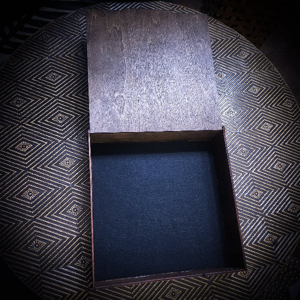 An open rectangular wooden box with a dark textured interior, photographed from above on a patterned black and gold surface.