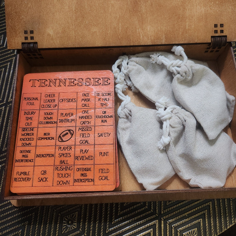 A Tennessee football-themed game set displayed inside a wooden box, featuring an orange square board with black printed text and a football icon, next to two small off-white drawstring pouches.