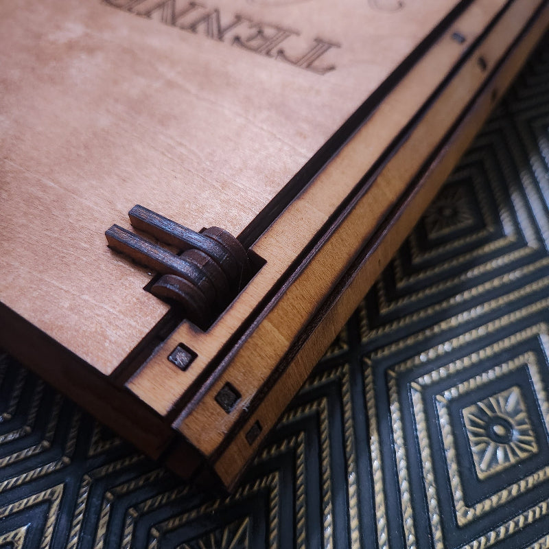 Close-up of a wooden box or case with a dark metal clasp, resting on a dark patterned surface. Engraved text is partially visible on the wood.