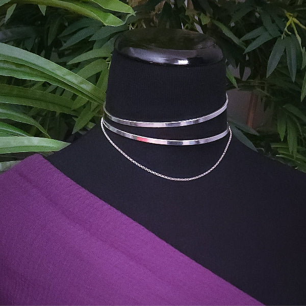 Silver asymmetrical collar with two bands and delicate chain. On a black mannequin with a purple fabric and green leaves in the background