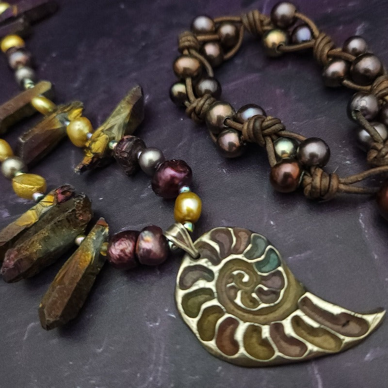 Make a bold statement with MARNIA, the one-of-a-kind necklace featuring titanium druzy, freshwater pearls, and a handcrafted bronze medallion ammonite. Turn heads with the vibrant colors and asymmetrical design, guaranteed to make you look and feel unforgettable!