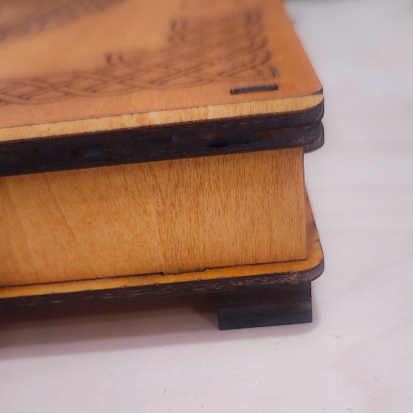 Close-up of a wooden box corner showing light natural wood grain on the lid and side, with dark stained edges and feet, photographed on a light neutral surface.
