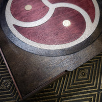 Close-up of a dark wooden box lid featuring a circular inlay with a red, silver, and gold triskelion symbol, resting on a patterned gold and black rug.