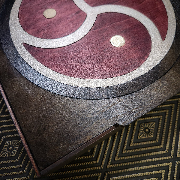 Close-up of a dark wooden box lid featuring a circular inlay with a red, silver, and gold triskelion symbol, resting on a patterned gold and black rug.