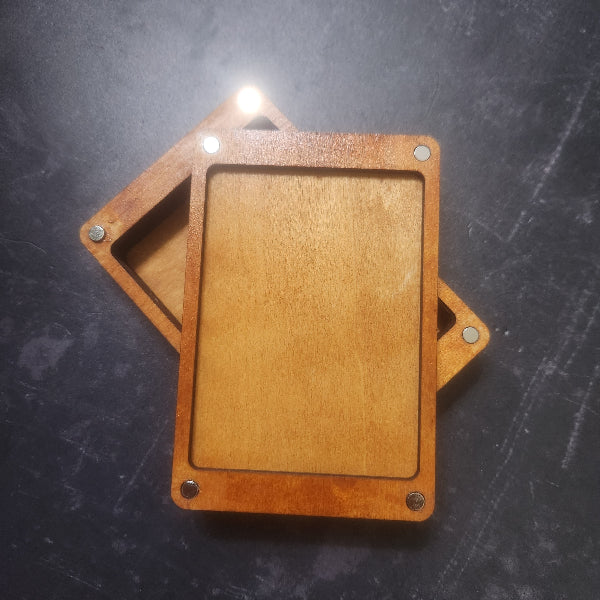 A wooden sliding card case with a light natural wood insert and medium brown outer frame, secured with metal corner screws, shown on a dark textured surface.