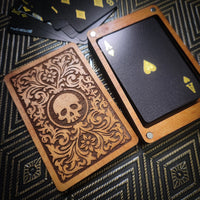 A deck of custom playing cards with a wooden case lid featuring a laser-engraved floral pattern and skull emblem, alongside a card showing a black back with gold Ace of Spades and a wooden border, set on a dark patterned surface.