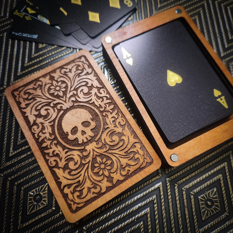 A deck of custom playing cards with a wooden case lid featuring a laser-engraved floral pattern and skull emblem, alongside a card showing a black back with gold Ace of Spades and a wooden border, set on a dark patterned surface.