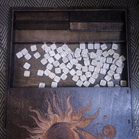An open wooden game box filled with many small square white tiles printed with symbols, photographed from above against a patterned dark background.