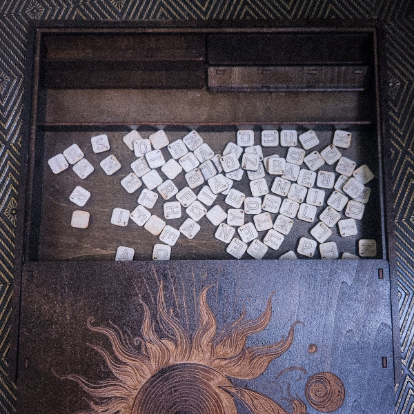 An open wooden game box filled with many small square white tiles printed with symbols, photographed from above against a patterned dark background.