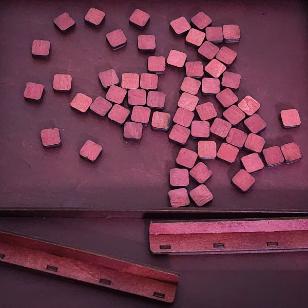 Small square wooden blocks with rounded corners scattered on a dark reddish-brown surface, with two matching wooden strips visible along the bottom edge.