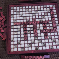 A vintage-style Scrabble board game set with a dark reddish-brown wooden grid, white square tiles, and pinkish-red letter tiles. The board is filled with words, and several loose red tiles are visible on the left side.