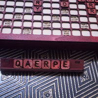 A wooden Scrabble-style word game board with red letter tiles spelling 'QAEERPE' on a patterned blue and white surface, with additional red tiles visible in the upper rack.
