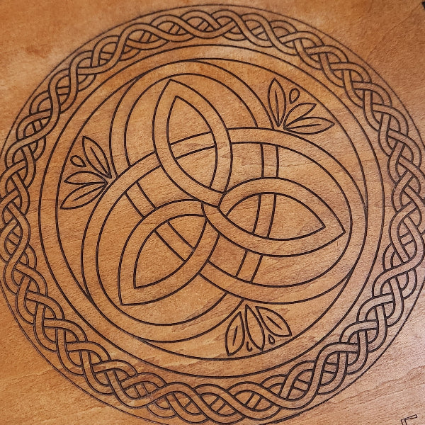 A circular Celtic knot design in dark brown line art on a warm, light brown wood-grain surface.