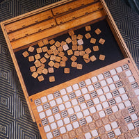 A wooden board game set with engraved letter tiles scattered on a black felt surface, photographed from above on a dark patterned fabric background.