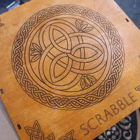 A close-up of a wooden Scrabble board showing a laser-engraved Celtic knot design and the word 'SCRABBLE' on a light natural wood surface.