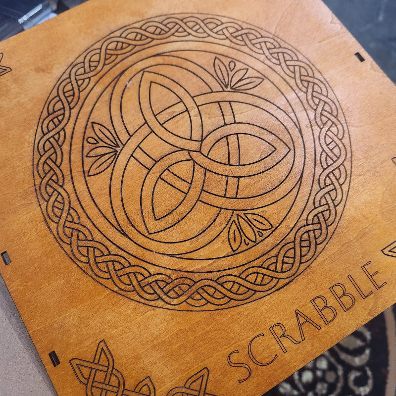 A close-up of a wooden Scrabble board showing a laser-engraved Celtic knot design and the word 'SCRABBLE' on a light natural wood surface.