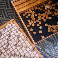 A wooden board game set shown in a styled flat-lay, featuring a wooden box with small square wooden tokens scattered inside, and a second wooden board with white square tiles printed with labels.