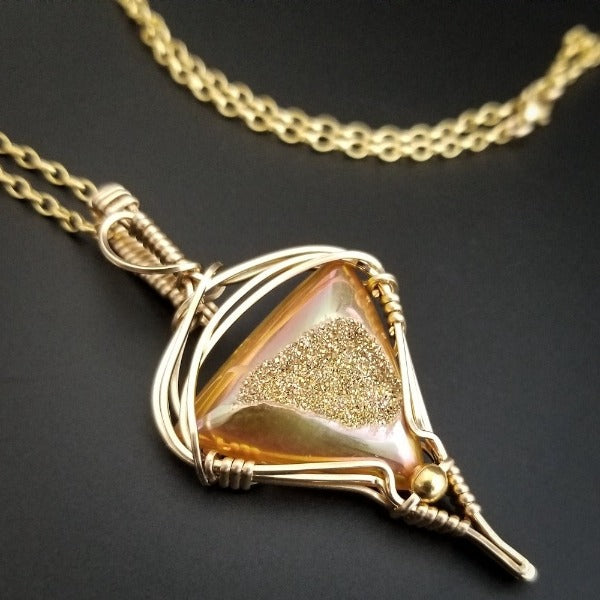 An incredibly luxurious look for a submissive collar. A triangular cut Gold Window Druzy is encased in a gold wire wrapped pendant that can be worn alone or slid onto most of our submissive collars. Featuring stunning open lattice work and a 14k gold filled chain that can be worn with a traditional clasp, or locked with a lock and key.